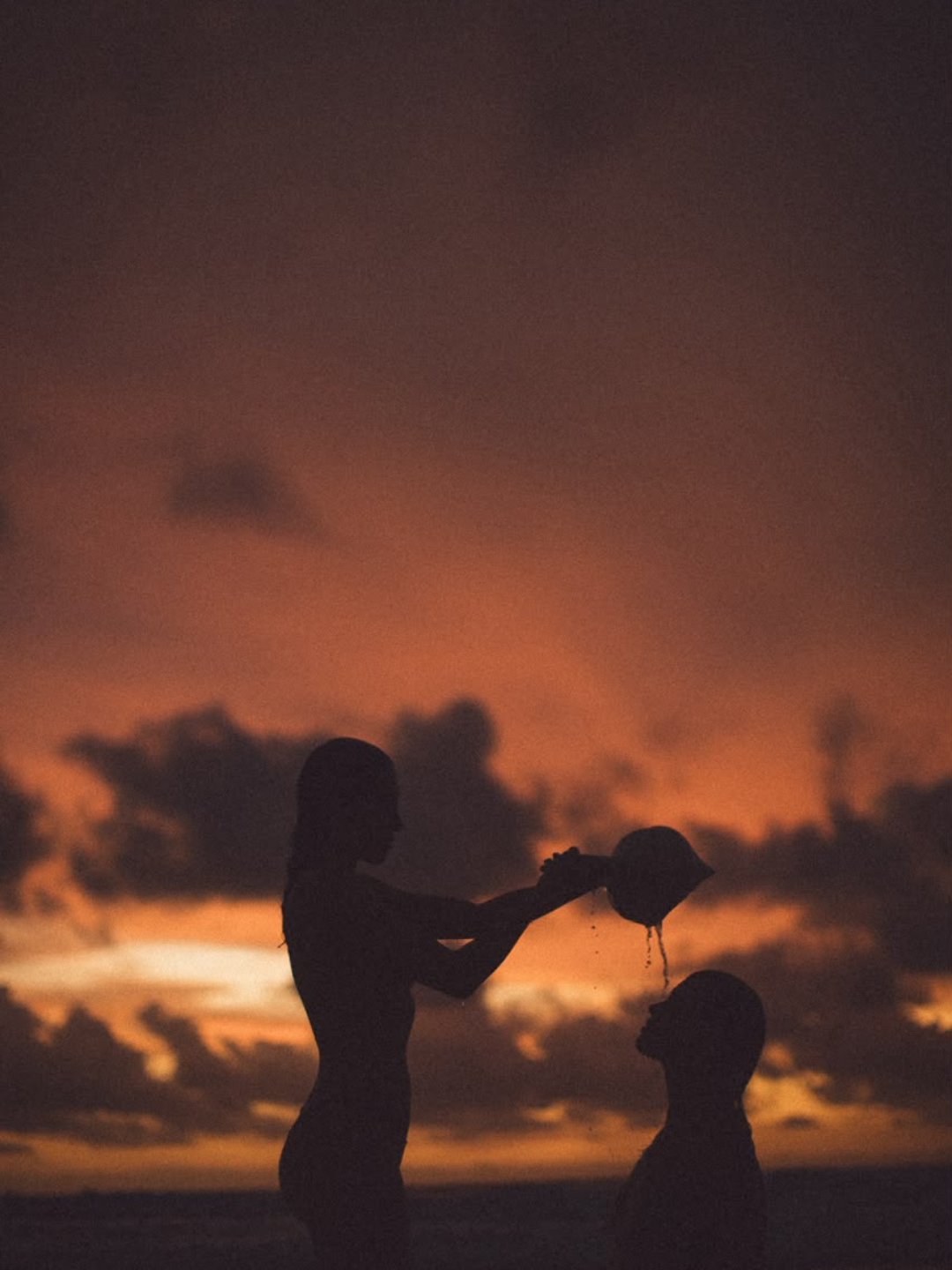 Two women in silhouette at sunset, one pouring water for the other
